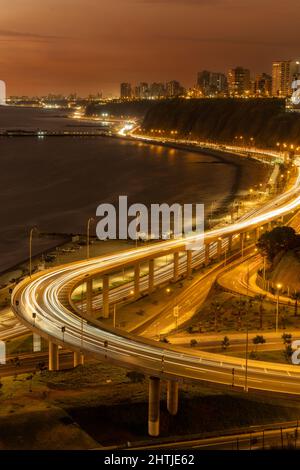 Lange Exposition der glühenden leeren Brücke in der Nähe des Meeres am Meer der Küstenstadt mit Wohngebäuden in der Nacht Stockfoto