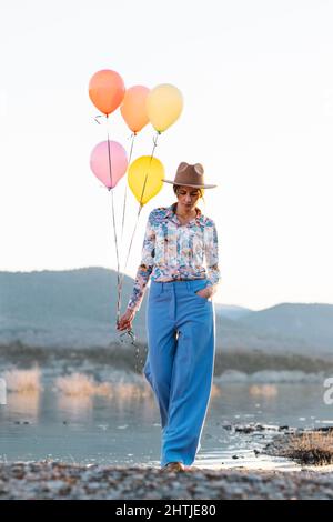 Nachdenkliche Hündin im Hut, die einen Haufen farbenfroher Ballons hält, während sie auf dem Ufer in der Nähe des plätschernden Sees steht Stockfoto