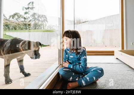 Seitenansicht des Mädchens im Pyjama, das mit süßem schwarzen amerikanischen Pit Bull Terrier spielt, während es in der Nähe einer Glastür auf dem Land mit grünem Rasen sitzt Stockfoto