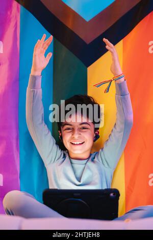 Aufgeregter arabischer Teenager-Junge in kabellosen Ohrhörern, der mit einem Tablet die Hände hochhebt und die Kamera gegen eine helle LGBT-Flagge anschaut Stockfoto