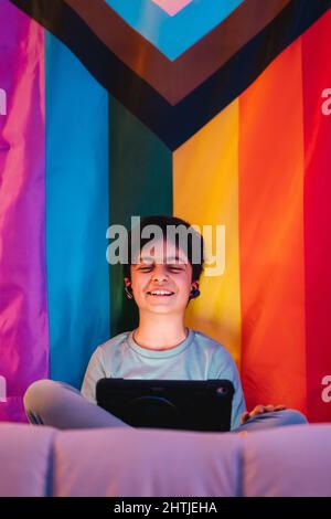 Niedriger Winkel des aufgeregten arabischen Teenagers mit geschlossenen Augen in kabellosen Ohrhörern, der ein Tablet mit fröhlichem Schrei gegen die helle LGBT-Flagge verwendet Stockfoto