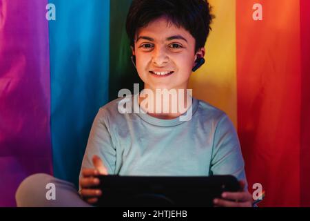 Aufgeregter arabischer Teenager-Junge in kabellosen Ohrhörern mit Tablet, das gegen eine helle LGBT-Flagge sitzt Stockfoto