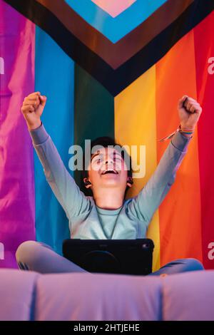 Ein aufgeregter arabischer Teenager mit geschlossenen Augen in kabellosen Ohrstöpseln, die mit einem Tablet die Hände hochheben, und einem freudigen Schrei gegen die helle LGBT-Flagge Stockfoto
