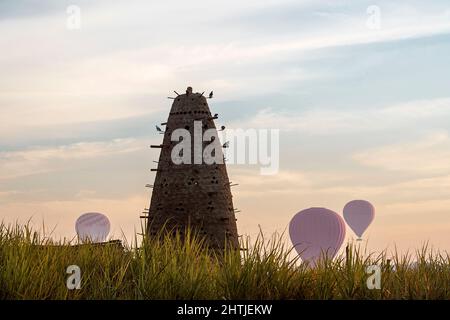 Hoher konischer Nesttaubenturm mit vielen Löchern auf grasbewachsenem Feld mit Luftballons in Ägypten am Sommertag Stockfoto