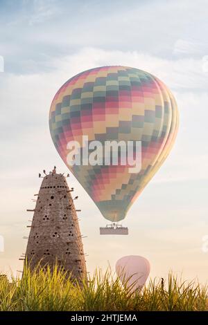 Hoher konischer Nesttaubenturm mit vielen Löchern auf grasbewachsenem Feld mit Luftballons in Ägypten am Sommertag Stockfoto