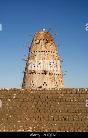 Hoher kegelförmiger ägyptischer Vogelnistturm mit Löchern und Holzstäben in der Nähe der Steinmauer gegen den wolkenlosen blauen Himmel in Ägypten Stockfoto