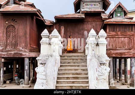 Mandalay, MYANMAR - 15. APRIL 2017: Seitenansicht eines kleinen asiatischen Mönchsjungen in orangefarbener Robe, der in der Nähe des alten hölzernen Shweinbin-Klosters in einem bewölkten Morgengrauen unterwegs ist Stockfoto