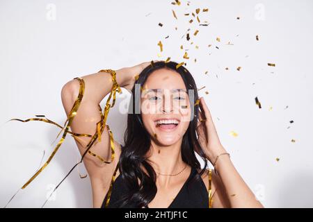 Positive junge Frau, die mit einem Lächeln auf die Kamera blickt, während sie während der Feiertagsfeier im Studio unter herabfallenden Konfetti auf weißem Hintergrund steht Stockfoto