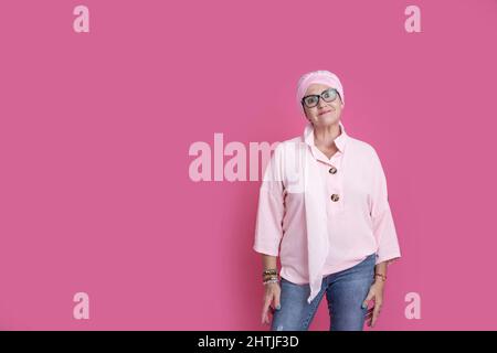 Positive reife Frau mit Krebs trägt Kopftuch und blickt mit einem Lächeln auf die Kamera, während sie im hellen Studio auf rosa Hintergrund steht Stockfoto