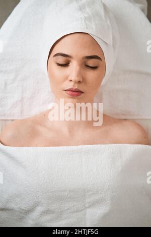 Ruhige weibliche Klientin, die mit geschlossenen Augen unter einer weißen Decke auf dem Massageliegen während einer Spa-Sitzung im modernen Schönheitssalon mit Vorräten liegt Stockfoto