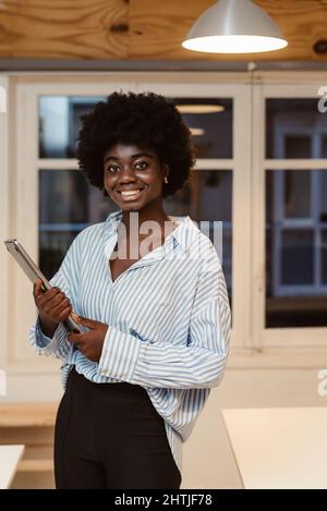 Fröhliche afroamerikanische Managerin in elegantem Outfit, die in einem hellen Büro mit Netbook in den Händen auf die Kamera schaut Stockfoto
