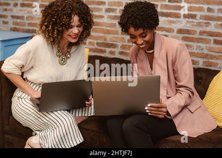 Positive multirassische weibliche Kollegen in elegantem Design stöbern in Netbooks, während sie gemeinsam auf der Couch im hellen, modernen Büro arbeiten Stockfoto