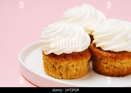 Köstliche Cupcakes aus Vanille mit süßem Schlagsahne-Zuckerguss serviert auf weißem runden Teller auf rosa Hintergrund in hellem Raum Stockfoto