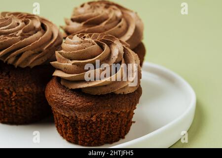 Leckere Cupcakes mit süßer Schokolade und köstlichem Schlagsahne-Zuckerguss, serviert auf einem runden weißen Teller auf grünem Hintergrund im Studio Stockfoto