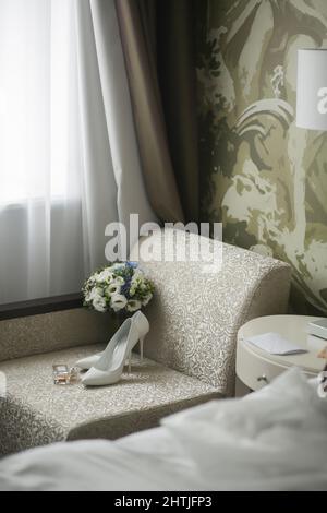 Weiße Hochzeitsblumen und elegante Schuhe mit Ringen in Glas-Box auf weichen Sessel in der Ecke des hellen Wohnung platziert Stockfoto