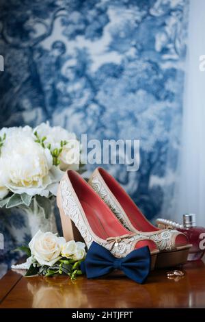Sanfte weiße Knospen und dekorierte Brautschuhe mit goldenen Ringen auf einem polierten Holztisch an der farbenfrohen Wand Stockfoto