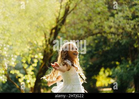 Glückliches junges Mädchen in Erstkommunion Kleid lächelnd und Blick auf weg, während im grünen Park an sonnigen Tag tanzen Stockfoto