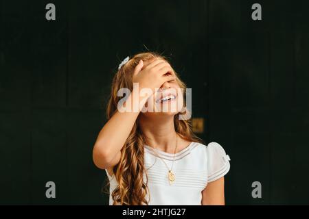 Positive junge Frau im Erstkommunionskleid mit langen Haaren und Hand auf Gesicht, die vor dunklem Hintergrund steht Stockfoto