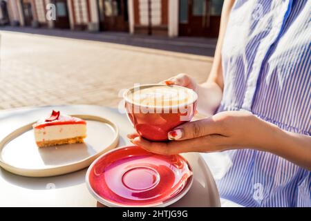 Tasse Latte-Kaffee in den Händen der Frau. Käsekuchen auf weißem Tisch draußen. Rote Tasse und roter Kuchen. Sonniger Tag. Stockfoto