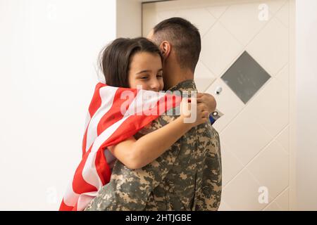 Glückliches kleines Mädchen Tochter mit amerikanischer Flagge umarmt Vater in Militäruniform kam aus der US-Armee zurück, männlicher Soldat wieder mit Familie zu Hause vereint Stockfoto