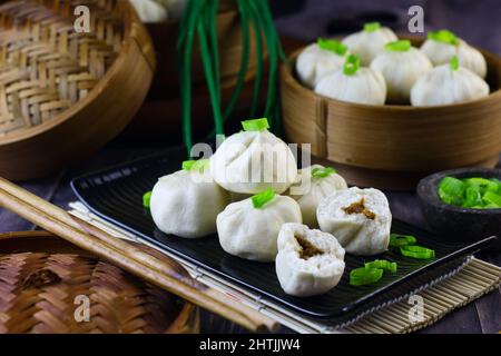 Bakpao Ayam oder Chicken Dampfgebäck auf schwarzem Keramikplatte serviert. Selektiver Fokus, dunkler Holzhintergrund. Stockfoto