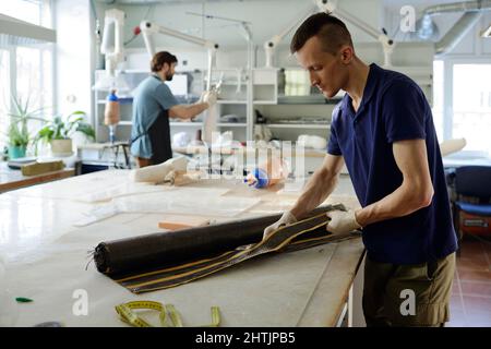 Junge Manufakturarbeiter mit Schere, die spezielles Material am Arbeitsplatz während des Prozesses der Herstellung von Beinprothesen schneidet Stockfoto