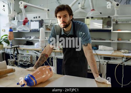 Junger selbstbewusster Arbeiter der Manufaktur in Handschuhen und Schürze, der in einer großen Werkstatt neben der Werkbank steht und die Kamera anschaut Stockfoto