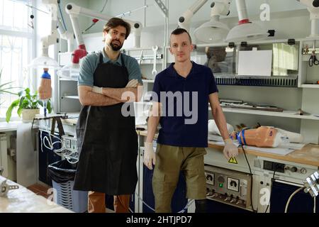 Zwei junge Spezialisten für Arbeitskleidung stehen in einer großen Werkstatt der prothetischen Manufaktur und schauen sich die Kamera an Stockfoto