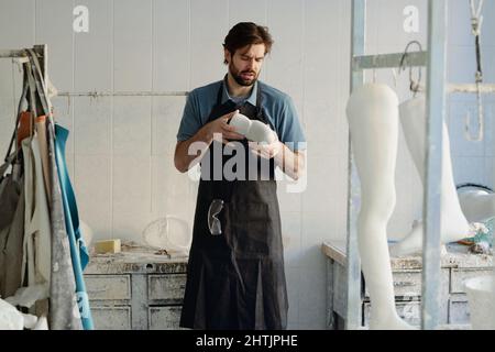 Der junge Meister der prothetischen Manufaktur blickt auf den Gipsguss in seinen Händen, während er gegen einen ausgestatteten Arbeitsplatz steht Stockfoto