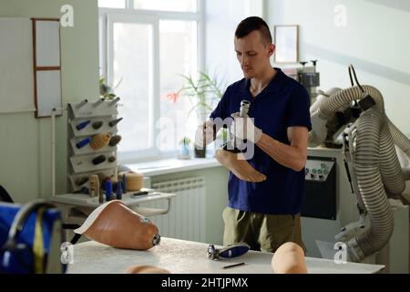 Junger Arbeiter in der Manufaktur, der Metallschaft in Kunststofffußguss fixiert, während er das ganze künstliche Glied am Arbeitsplatz zusammenführt Stockfoto