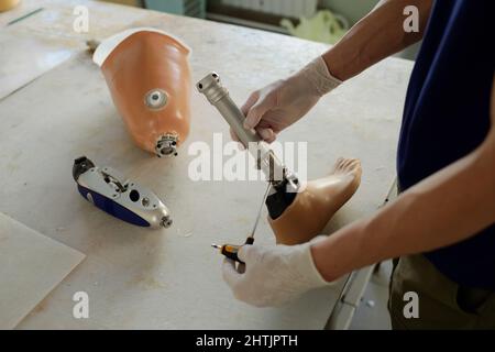 Hände von Handschuhen Handarbeiter der Fabrik hält Schraubendreher und Teil der Beinprothese über dem Tisch, während die Stahlwelle zu befestigen Stockfoto