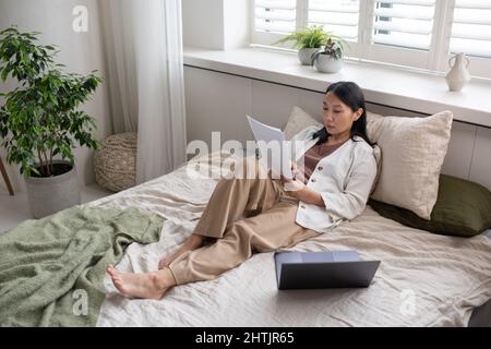 Junge, selbstbewusste asiatische Frau in legerer Kleidung, die durch die Finanzpapiere schaut, während sie auf dem Doppelbett vor einem großen Fenster sitzt Stockfoto