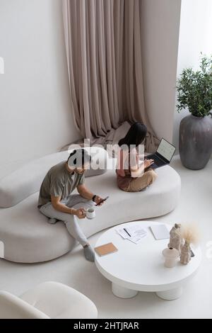 Junge asiatische Geschäftsfrau bereitet Präsentation vor, während Mann in Casualwear neben ihrem SMS auf dem Smartphone sitzt Stockfoto