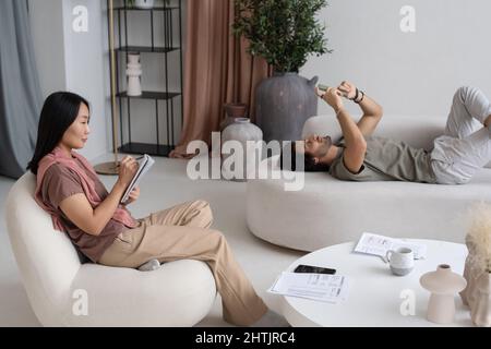Die junge asiatische Frau schreibt den Arbeitsplan auf die Seite des Notizbuchs, während ihr kaukasischer männlicher Mitarbeiter im Mobiltelefon schreibt Stockfoto