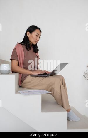 Junge seriöse asiatische Frau in Pastellfarben Casualwear sitzt auf weißem Treppenhaus und bereitet die Präsentation des Business-Projekts Stockfoto