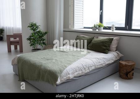 Doppelbett mit weißen und baumgrünen Kissen und grauem karierten Kissen, die an einem großen Fenster stehen, in einem geräumigen Schlafzimmer Stockfoto