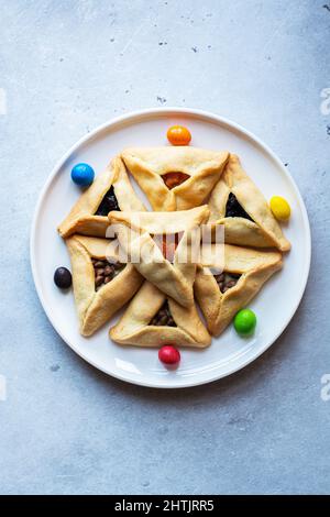 Traditionelle Kekse für den jüdischen Feiertag Purim - Hamantashen mit verschiedenen Füllungen und bunten Bonbons auf einem Teller. Festliches Backkonzept. Stockfoto