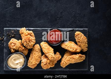 Gebratenes Hähnchenflügel im Kentucky-Stil, knuspriges Huhn serviert mit Saucen auf schwarzem Schiefergrund. Kopiespeicherplatz in der Draufsicht Stockfoto