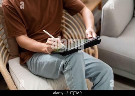 Technologiekonzept der junge Mann, der auf dem Stuhl sitzt und sein iPad hält und etwas darauf notiert. Stockfoto