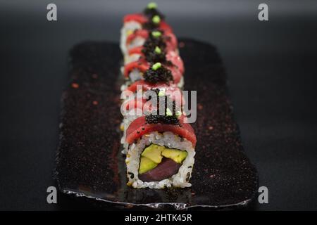 Bunte Kombination aus traditionellem japanischen Uramaki Sushi-Rolle mit Thunfisch, Avocado, schwarzem Kaviar und Wasabi auf der Oberseite. Nahaufnahme auf schwarzem Hintergrund. Stockfoto