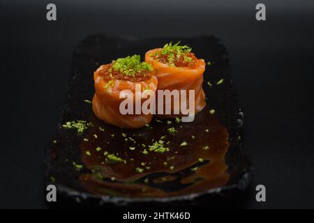 Traditioneller japanischer Lachs-Gunkan, zwei Stücke Wildlachsfilet, frisch geriebene Limette und Lachskaviar auf schwarzem Teller mit schwarzem Hintergrund. Stockfoto