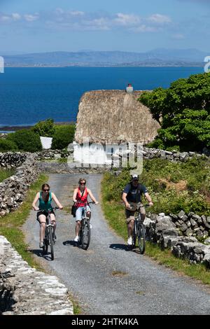 Weiß gewaschene Hütte mit Strohgedeckten Radfahrern zwischen Trockenmauern und Galway Bay dahinter, Inishmore, Aran Islands, County Galway, Irland Stockfoto