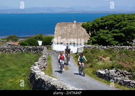 Weiß gewaschene Hütte mit Strohgedeckten Radfahrern zwischen Trockenmauern und Galway Bay dahinter, Inishmore, Aran Islands, County Galway, Irland Stockfoto