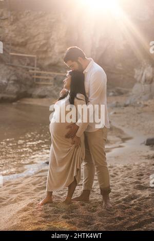 Ein trächtiges Paar genießt den Sonnenuntergang am Strand. Schwangere blicken in den Horizont, während ihr Mann sie küsst. Stockfoto