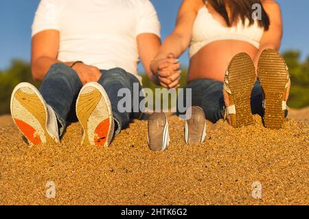 Ein Schwangeres Paar sitzt am Strand und hält an einem sonnigen Tag die Hände bereit, Eltern zu werden. Stockfoto