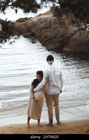 Schwanger junge Frauen lächeln und umarmen ihren Mann am Strand, der Mann hält ihren schwanger Bauch und blickt zum Horizont. Stockfoto