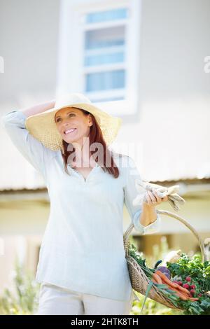 Im Garten zu arbeiten ist, wie ich mich erhole. Eine schöne Frau hält ihre Gartenhandschuhe und einen Korb mit frisch gepflücktem Gemüse, während sie in ihrem Garten steht. Stockfoto