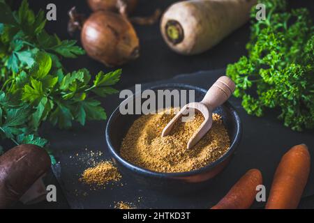 Hausgemachte Gemüsebrühe Pulver in einer schwarzen Schüssel, Bio-Gemüsebrühe, mit rohem Gemüse auf schwarzem Hintergrund Stockfoto
