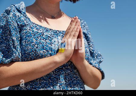 Ukrainische Frau mit einem Flaggensymbol auf der Hand betet gegen den blauen Himmel. Krieg beenden Stockfoto