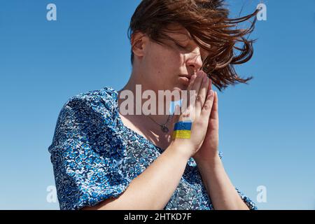 Ukrainische Frau mit einem Flaggensymbol auf der Hand betet gegen den blauen Himmel. Krieg beenden Stockfoto